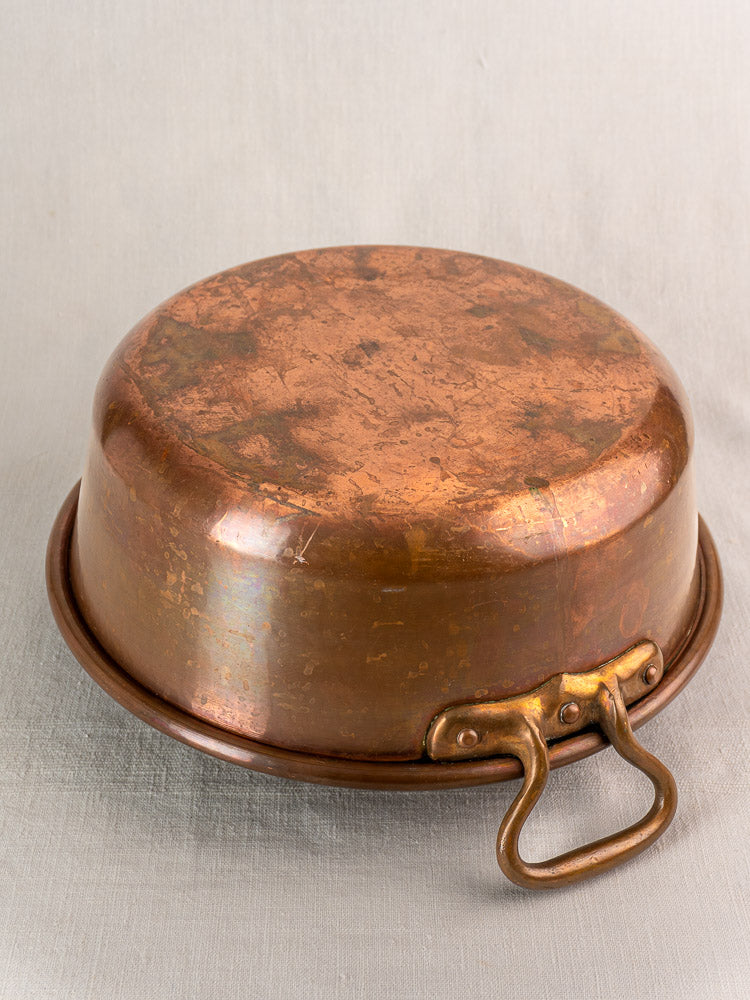 Antique French copper basin upside down, showing aged brass handle on light gray fabric backdrop.
