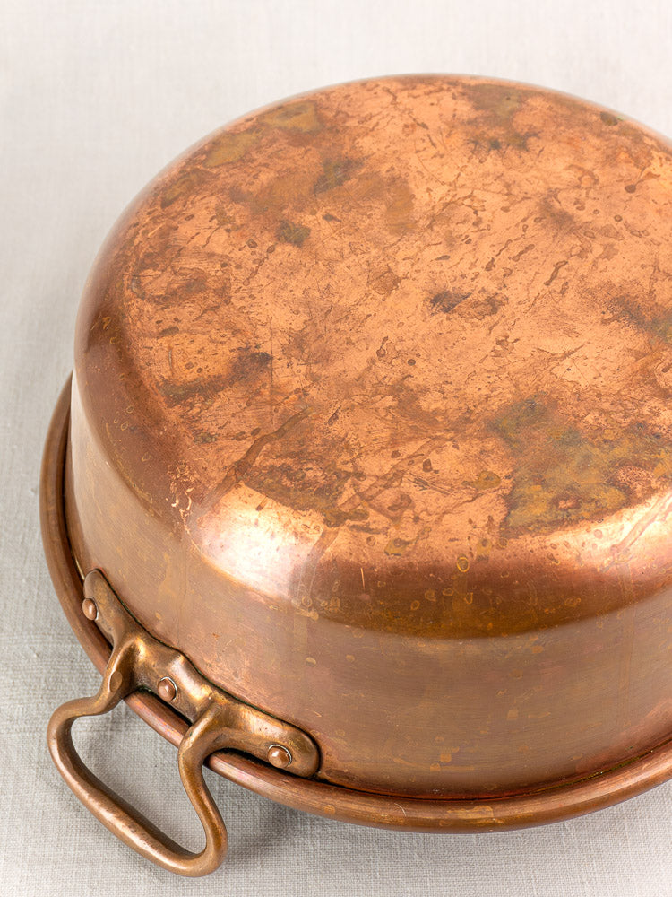Antique French copper basins with worn bottoms and brass handles, nesting on a light cloth background.