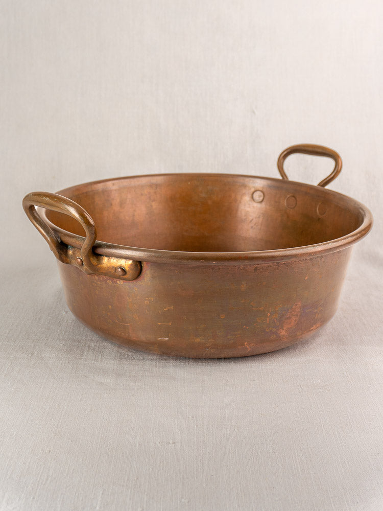 Antique French copper basin with two curved handles and rustic patina, displayed on a light surface.