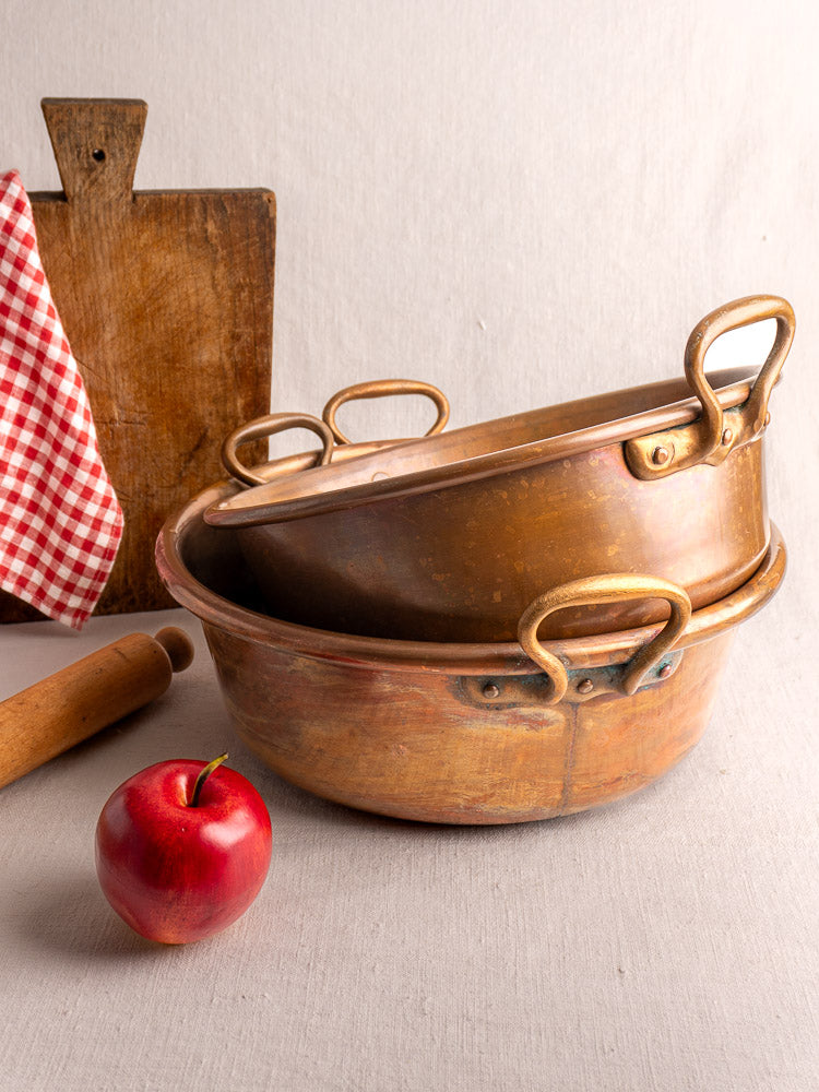 Pair of late 19th century French copper basins with brass handles, nested beside a red apple and checkered cloth.