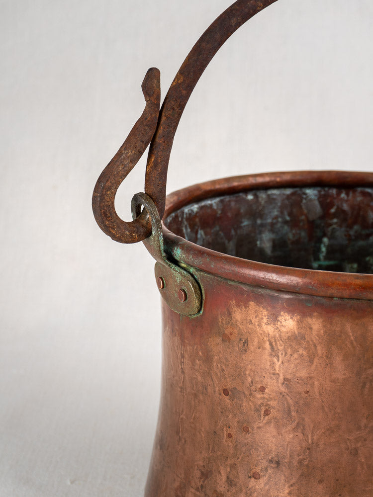 Antique French hammered copper bucket with green patina and wrought iron handle on light background.
