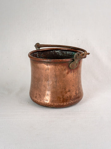 Antique French hammered copper bucket with wrought iron handle on a light fabric background.
