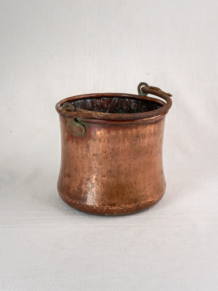 Antique French hammered copper bucket with wrought iron swing handle on light fabric surface.