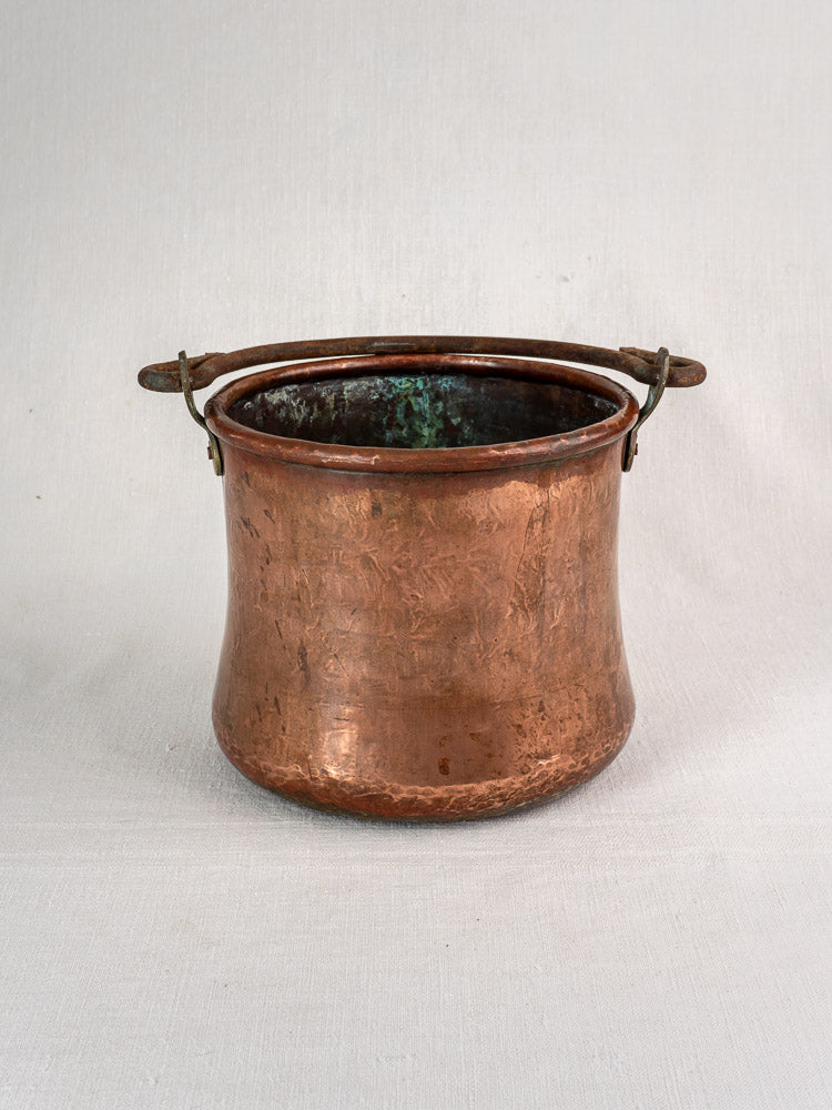 Antique French hammered copper bucket with green interior patina and wrought iron handle on fabric background.