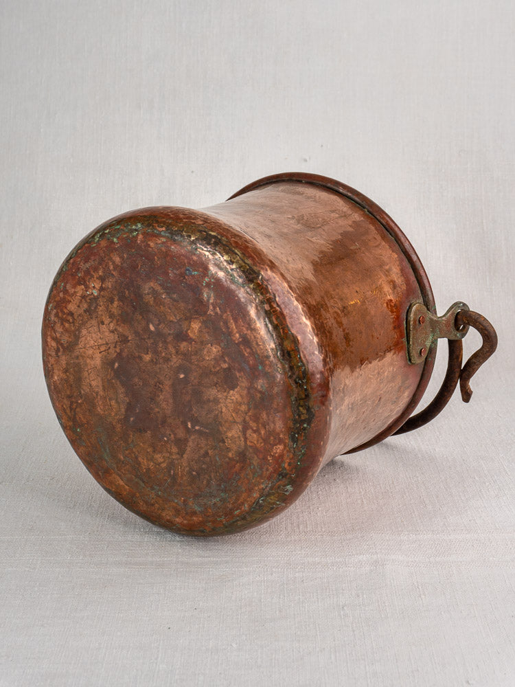 Antique French hammered copper bucket with wrought iron swing handle on light gray textured surface.