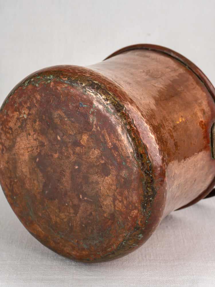 Antique French hammered copper bucket with aged patina and iron swing handle on fabric surface.