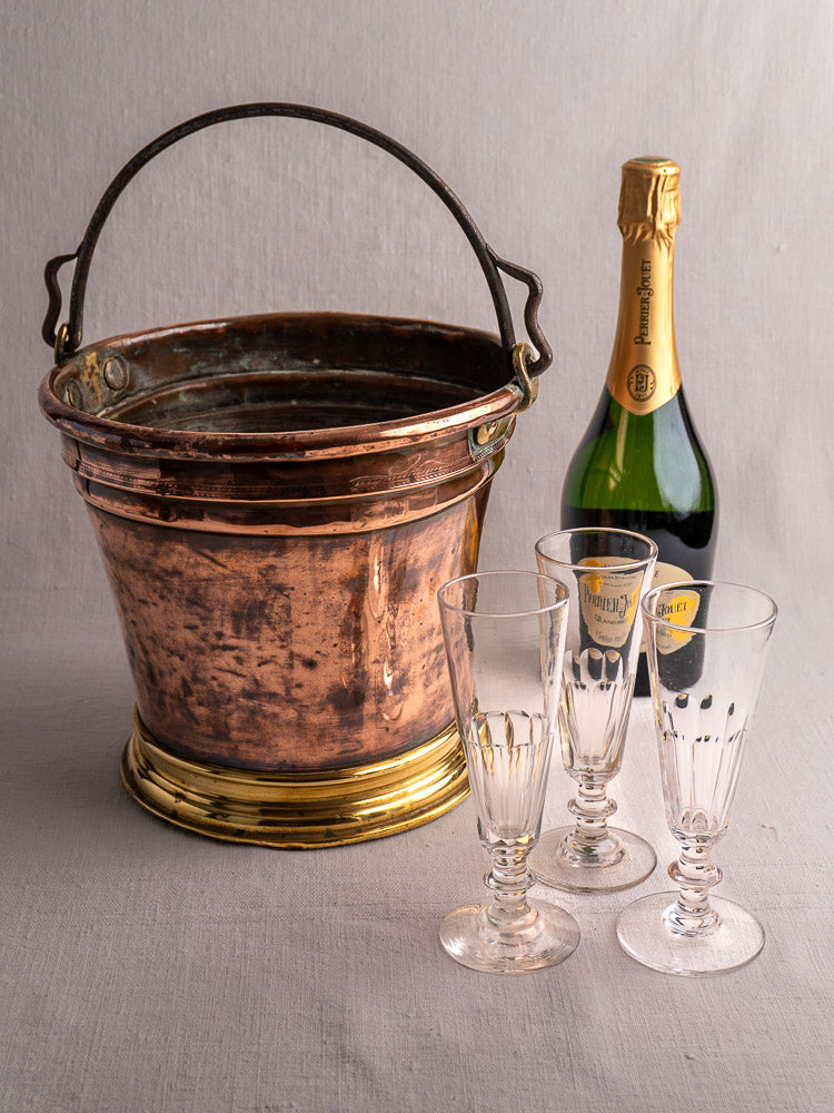 Antique French copper champagne bucket beside a bottle and three flutes on a light gray surface.