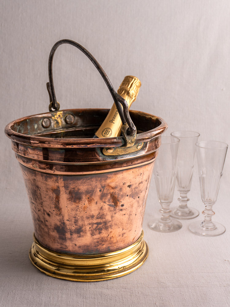 Antique French copper champagne bucket with bottle, beside three empty crystal glasses on a pale tablecloth.