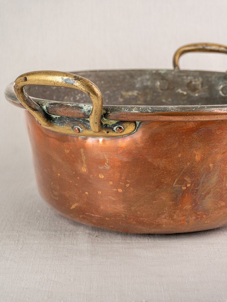 Antique French copper jam pan with brass handles and rich patina on a neutral fabric surface.