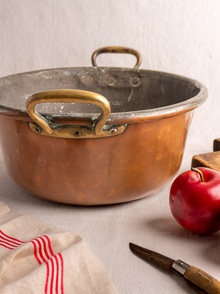 Antique French copper jam pan with brass handles and worn patina on a neutral background.