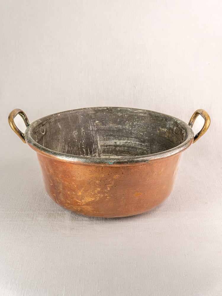 Antique French copper jam pan with brass handles and worn patina on a neutral background.