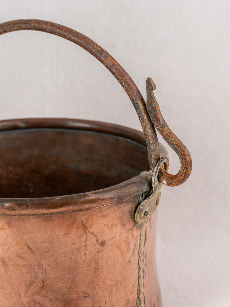 Antique French copper cauldron with hammered finish and iron bail handle on a plain light fabric background.