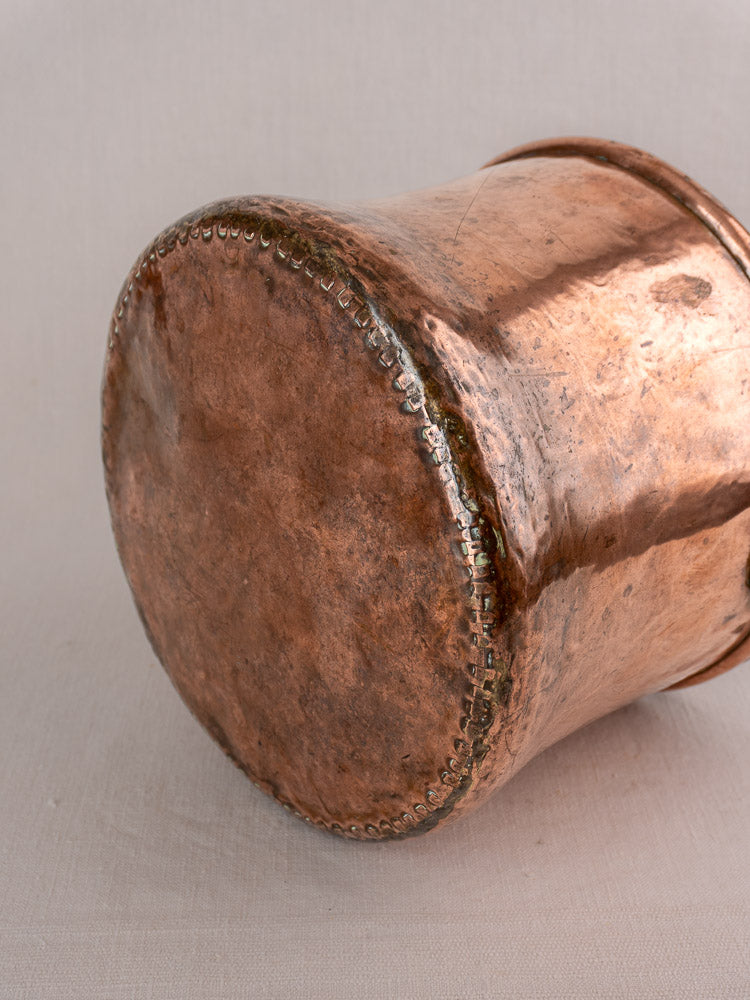 Antique French hammered copper cauldron with iron bail handle, visible seams and textured base, on light fabric.