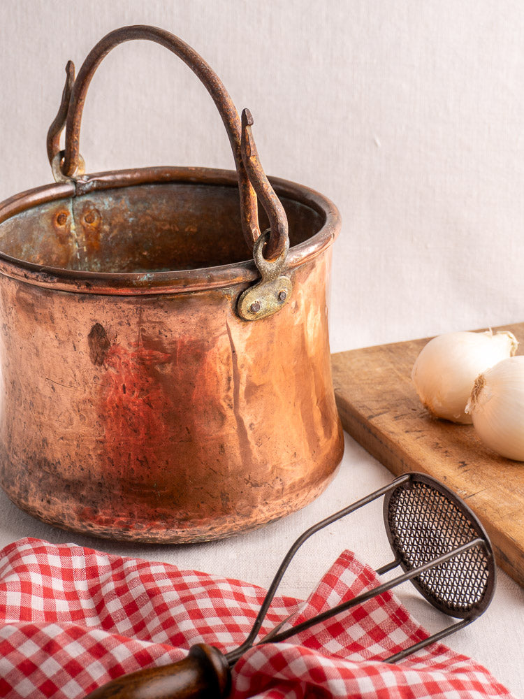 Antique French copper cauldron with hammered finish and iron handle, styled with onions and vintage kitchen accents.