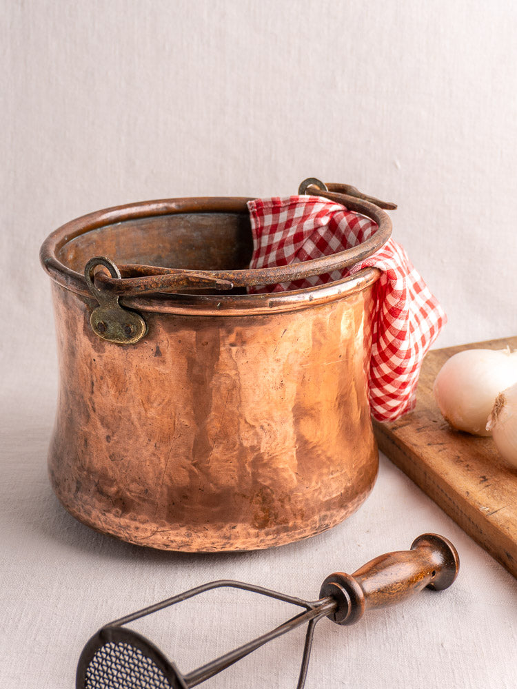 Antique French copper cauldron with hammered finish and iron handle, on white surface with checkered cloth and kitchen props.