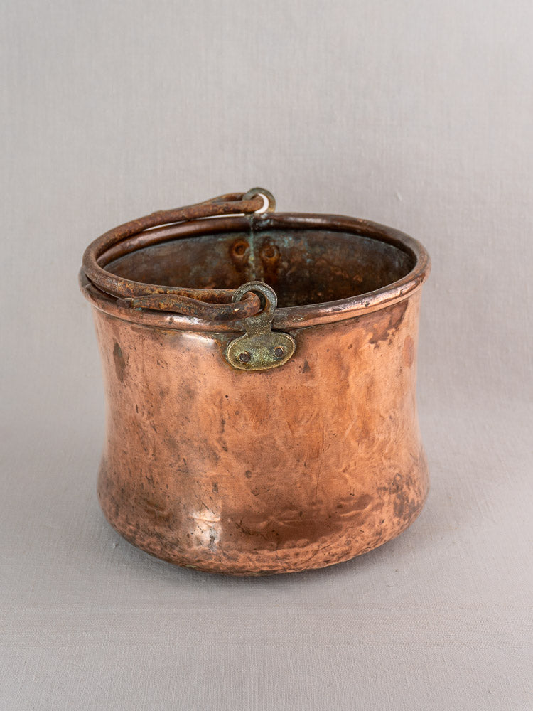 Antique French copper cauldron with hammered texture, iron bail handle, rich patina, on light gray surface.