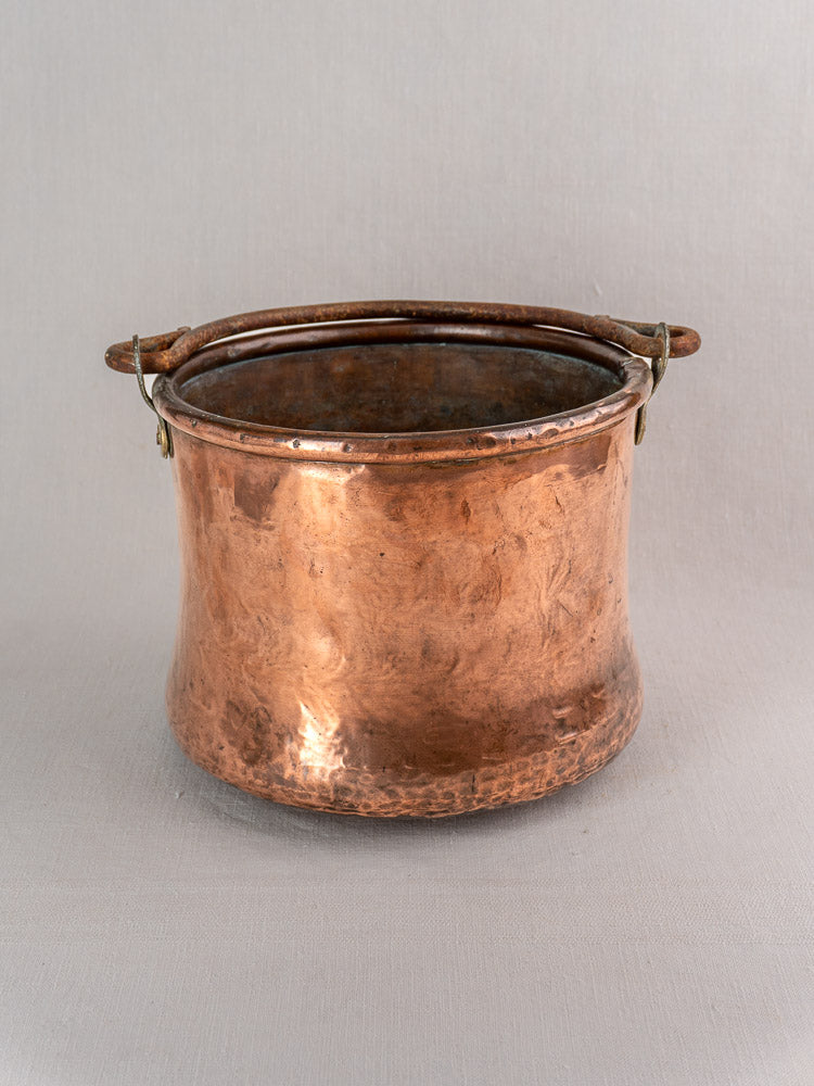Antique French copper cauldron with hammered finish and iron bail handle, flared rim, on a plain gray background.