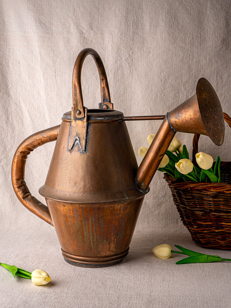 Vintage French copper watering can by Jean Goardère, Villedieu MOF, beside yellow tulips in a woven basket.