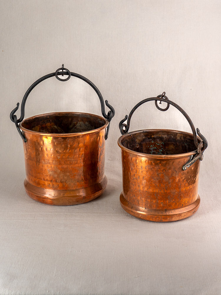 Pair of 19th-century French antique copper hanging cauldrons with dark metal handles on a light neutral surface.