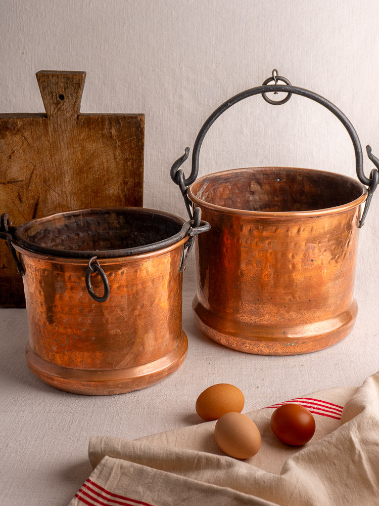 Pair of 19th-century French copper hanging buckets on a light surface with a cutting board, brown eggs, and red-striped cloth.