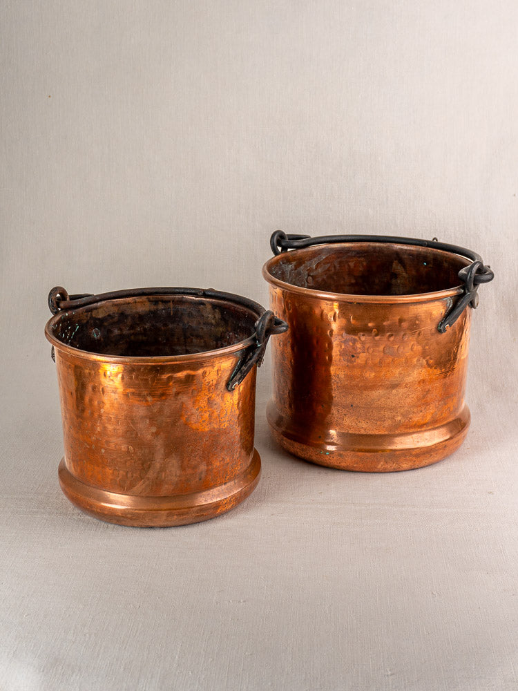 Pair of 19th-century French copper hanging buckets with dark metal handles and hammered finish on beige surface.