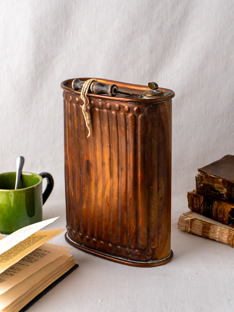 French copper bouillotte vintage fluted hot water bottle on table with books and green mug.