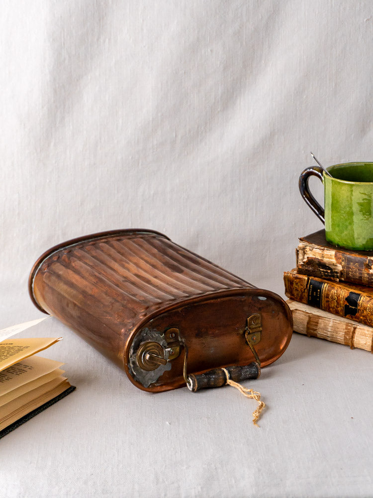 French copper bouillotte hot water bottle with handle and key beside old books and green mug.