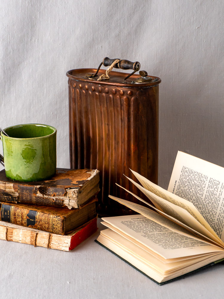 French copper bouillotte and green mug displayed with vintage books on neutral fabric backdrop.