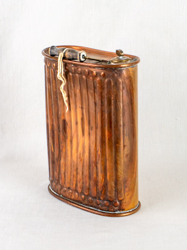 Antique French fluted copper bouillotte hot water bottle with wooden handle on pale background.