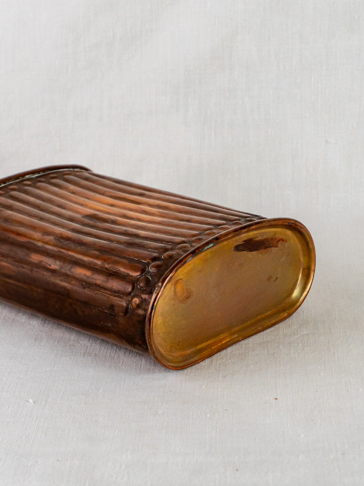 Vintage French copper bouillotte hot water bottle with fluted ribbing and rounded ends on light gray fabric.