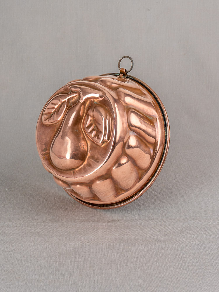 19th-century antique French copper jelly mold with embossed pear motif, fluted edge, and hanging loop on gray surface.