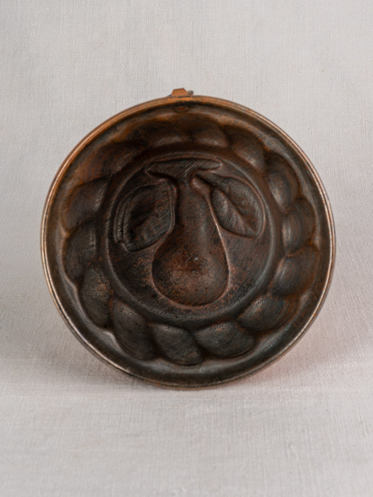 Antique French copper jelly mold, round with embossed pear and leaves, scalloped border, on a light fabric background.