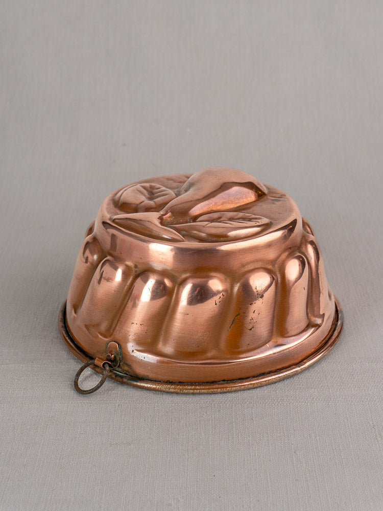 19th-century antique French copper jelly mold with embossed pear motif and side ring, set against a neutral gray background.