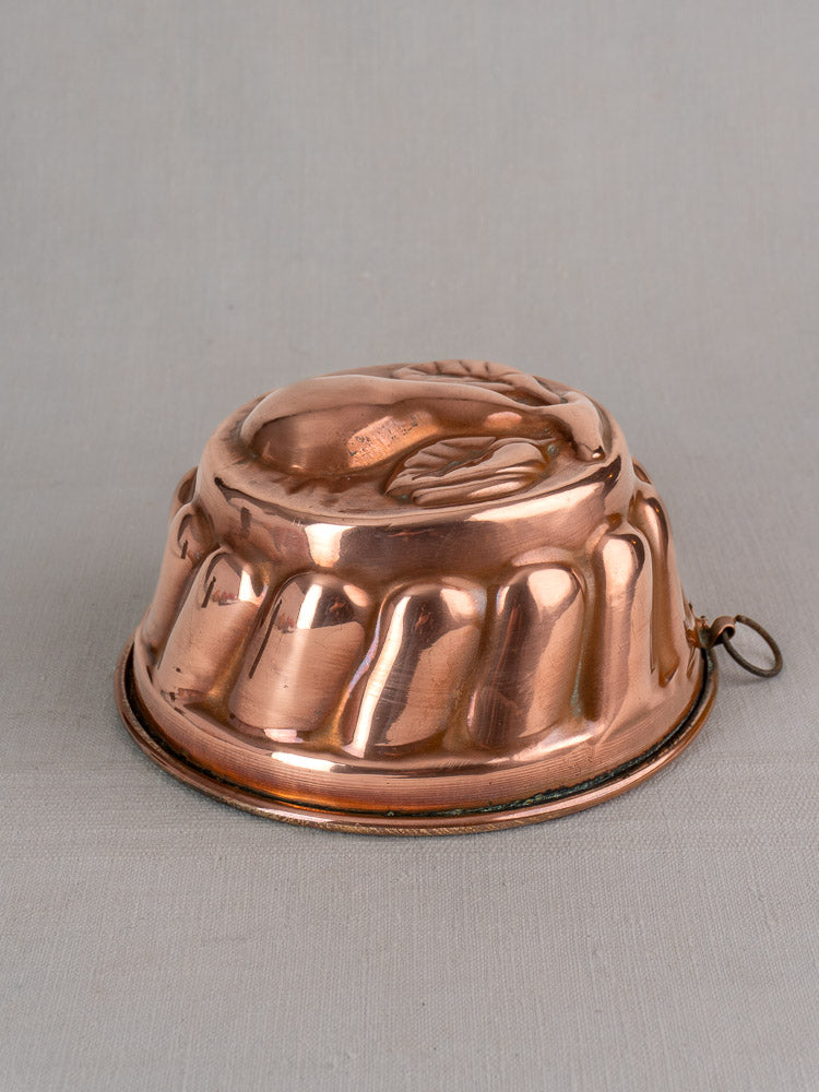 Antique French copper jelly mold with scalloped edge and embossed pear, side ring for hanging, on neutral fabric.