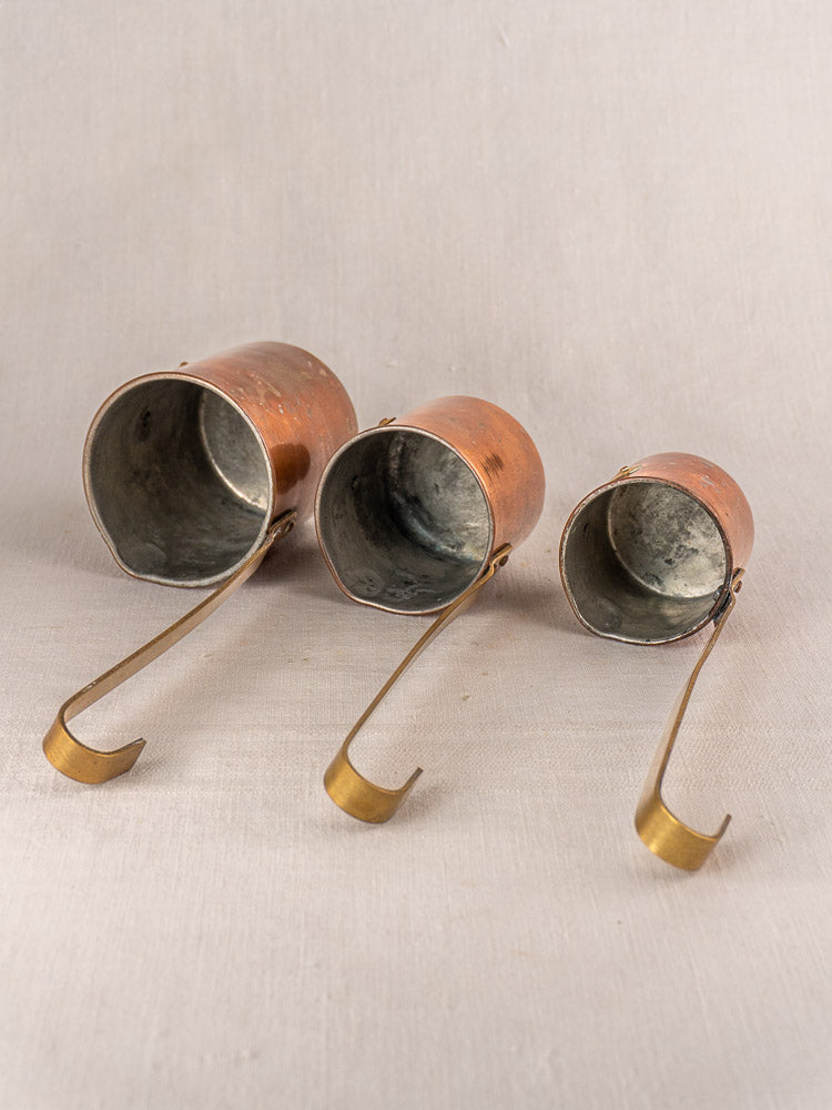 Set of 5 antique French copper mesures with brass handles on a neutral fabric background.