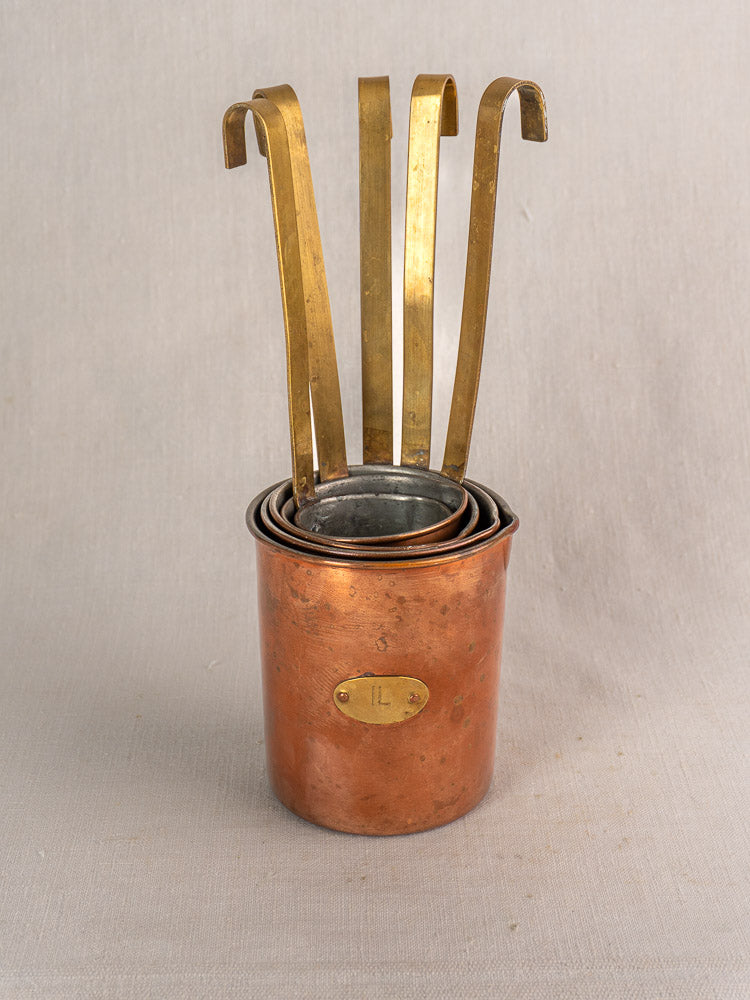 Set of five late 19th-century French copper measures with brass handles and labels on neutral fabric background.