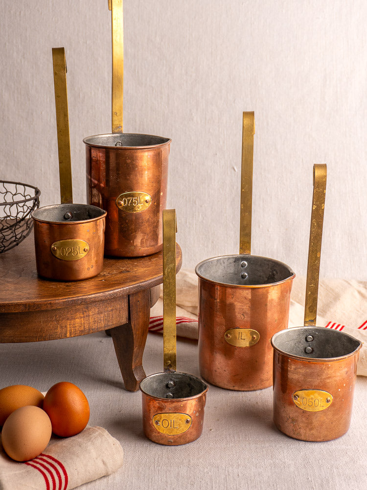 Set of five antique French copper measuring cups with brass handles and volume labels, elegantly arranged.