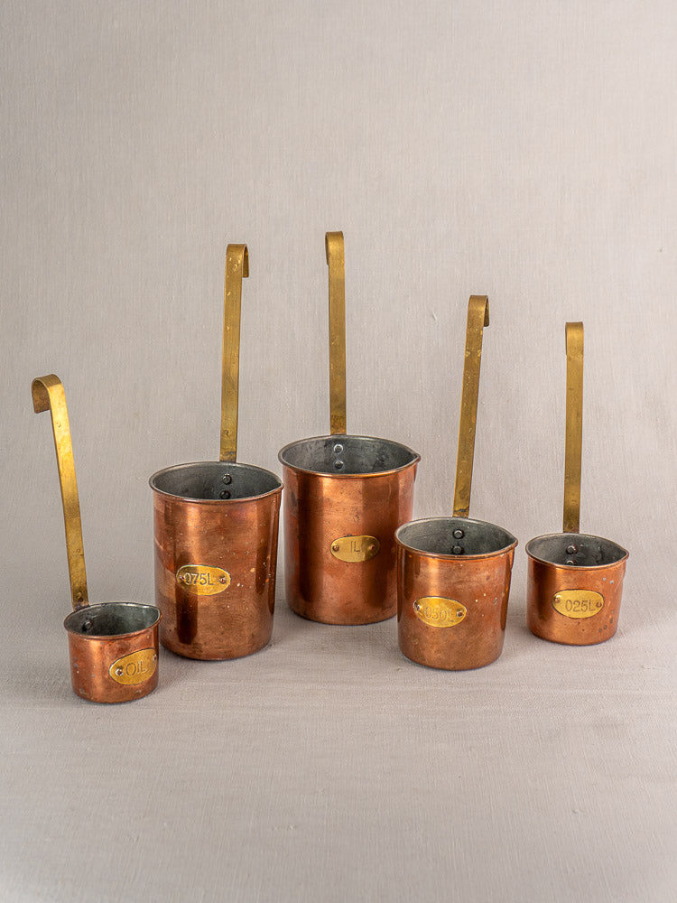 Set of five antique French copper measuring cups with brass handles and volume labels, elegantly arranged.