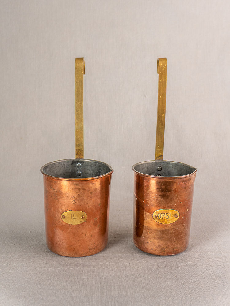 Set of five late 19th-century French copper measure cups with brass handles and labels, arranged side by side.
