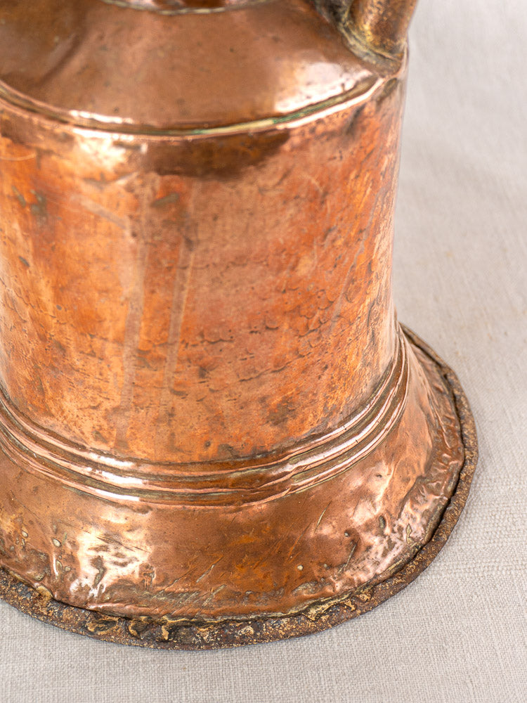 Antique French hammered copper water jar with twin iron handles and wide base, aged patina, on light fabric.