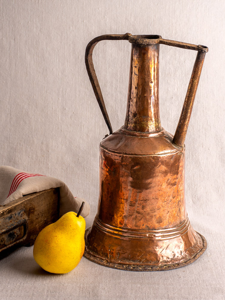 Antique French copper water jar with twin iron handles on beige cloth beside a yellow pear and striped fabric-covered box.