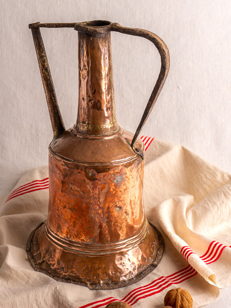 19th-century French copper water jar with hammered finish and twin iron handles on cream cloth with red stripes.
