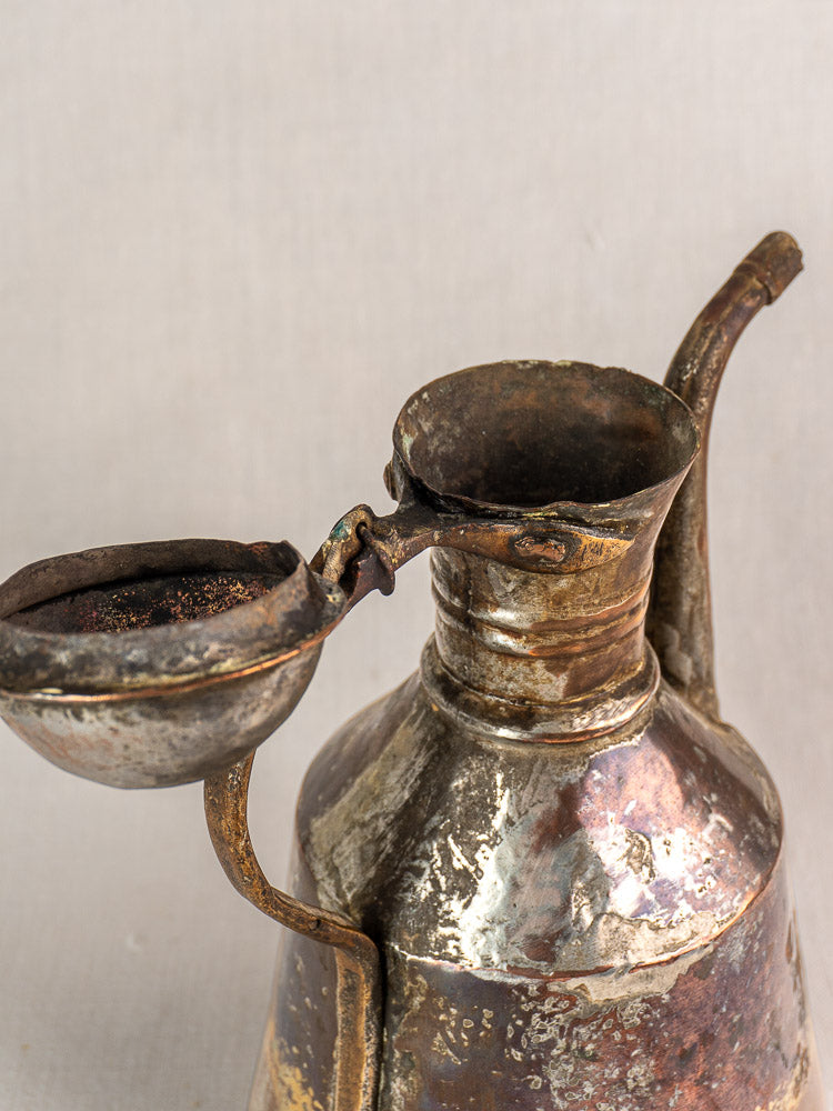 19th century antique French copper oil pouring jug, hand hammered with curved spout, lid, and handle; worn patina. Neutral fabric backdrop.