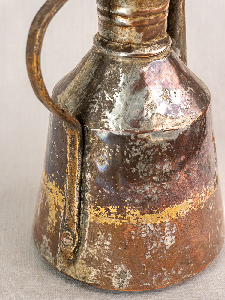 Antique French copper oil pouring jug, hand hammered with handle, tarnish, rust, and patina on a neutral background.