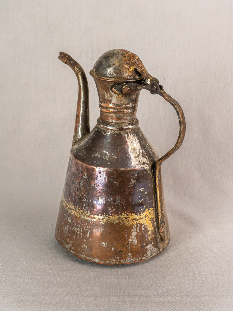 19th century antique French copper oil pouring jug, hand hammered with curved spout, lid, and handle; worn patina. Neutral fabric backdrop.