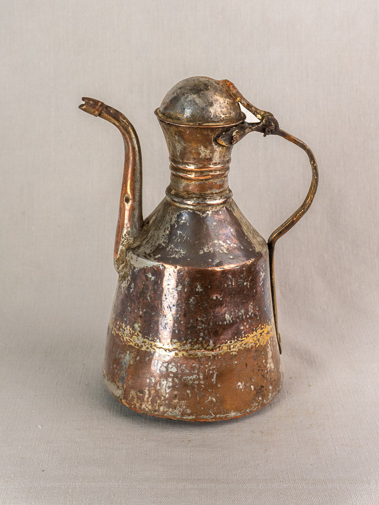 Antique French copper oil pouring jug with long spout, rounded lid, and curved handle on a light fabric background.