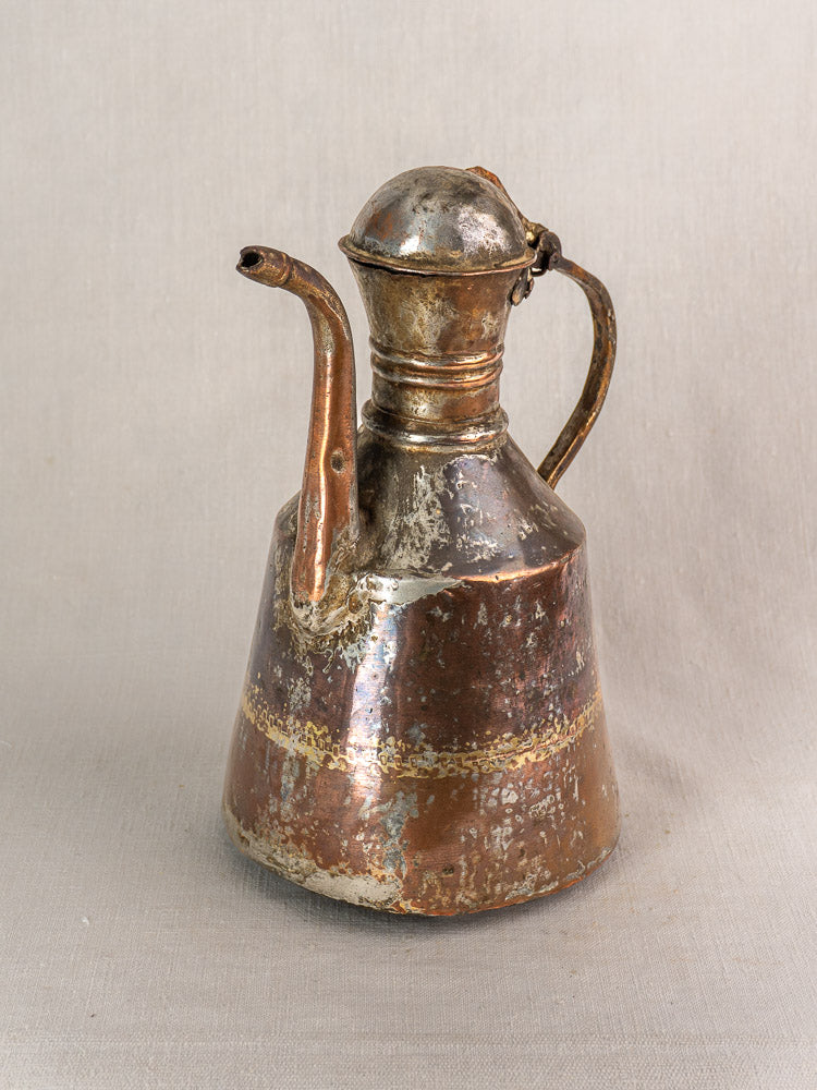 Antique French hand-hammered copper oil pouring jug with domed lid, curved spout, and worn patina on neutral fabric.