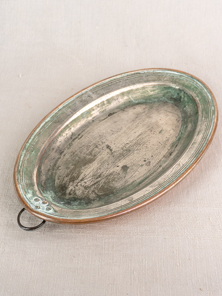 Antique French copper platter with tinned oval form and hanging ring, rich patina, on light fabric background.