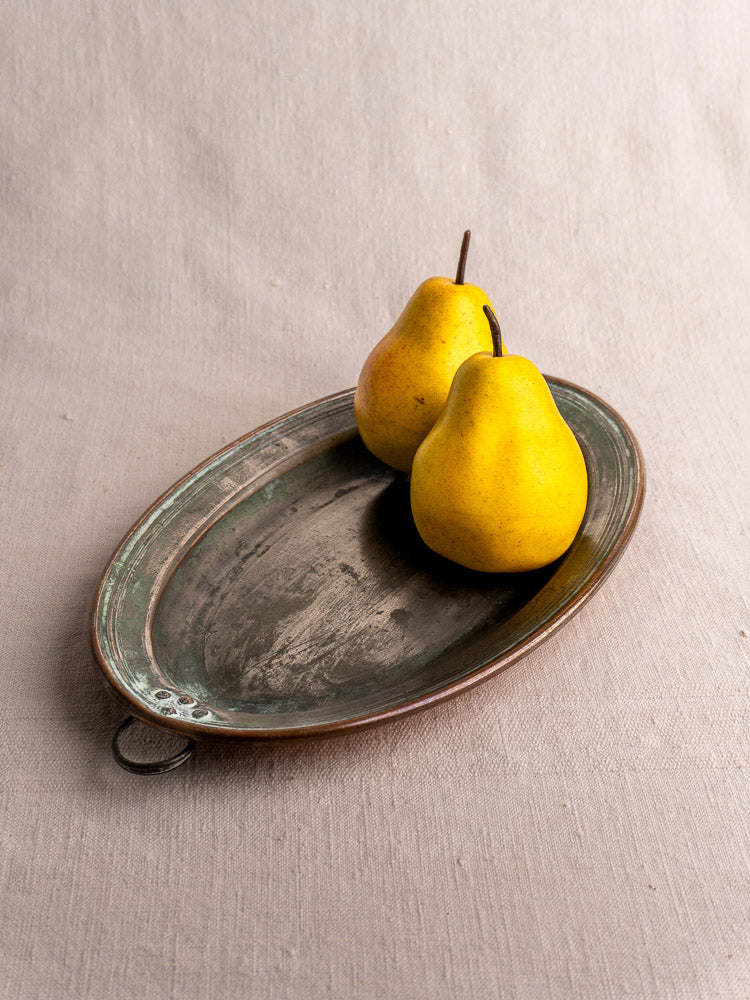 Antique French copper platter with hanging ring, tinned oval dish shown atop beige fabric with two yellow pears.