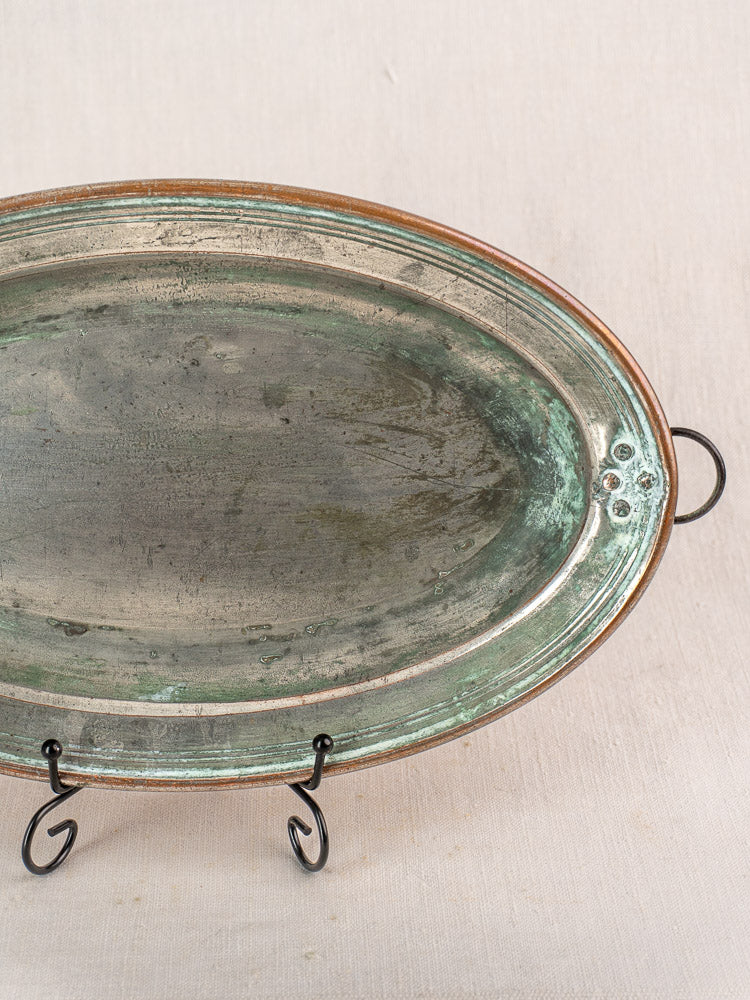 Antique French copper oval platter with hanging ring, tinned interior, weathered patina on black metal stand against neutral fabric.